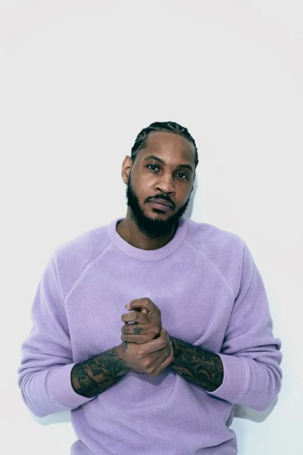 Portrait of Black man with braided hair indoors against white wall. Man wears lavender crewneck sweatshirt and poses with hands clasped.