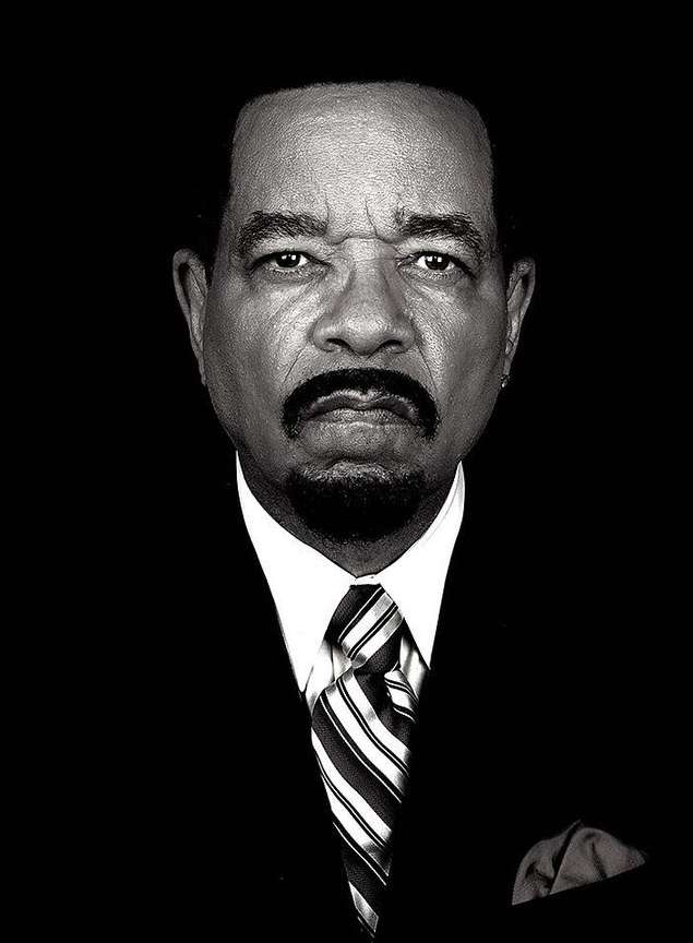 Black and white portrait of Ice-T in a suit and tie looking at the camera against a dark background in a medium shot, head and shoulders.