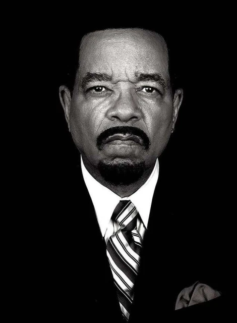 Black and white portrait of Ice-T in a suit and tie looking at the camera against a dark background in a medium shot, head and shoulders.