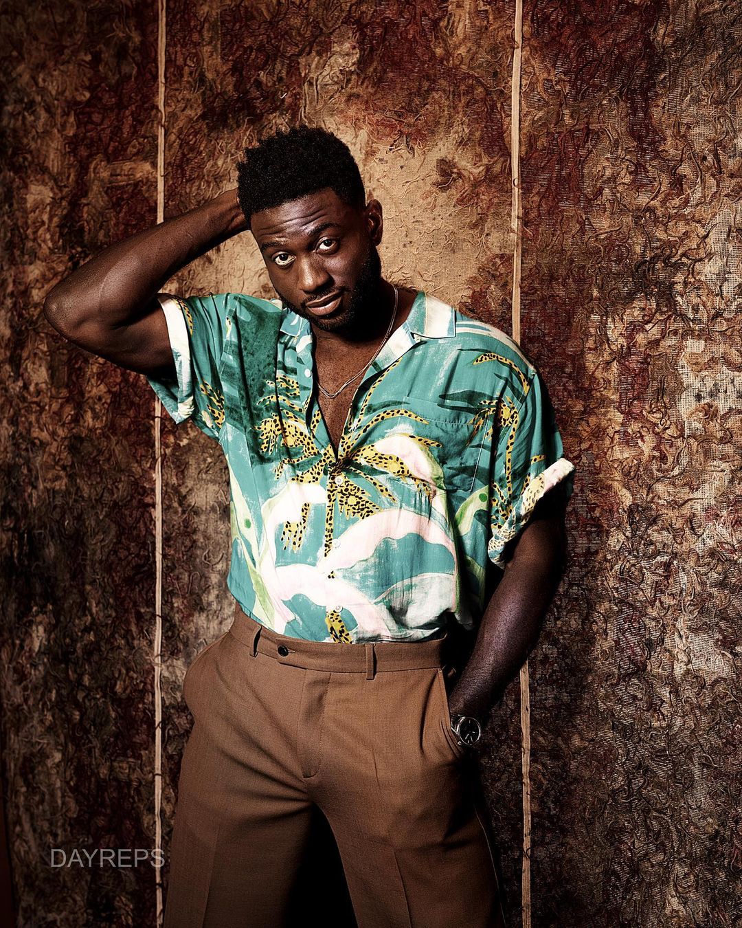 A stylish man with a hand behind his head poses indoors wearing a patterned short-sleeve shirt and brown pants against a mottled backdrop.