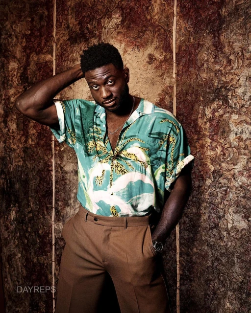 A stylish man with a hand behind his head poses indoors wearing a patterned short-sleeve shirt and brown pants against a mottled backdrop.