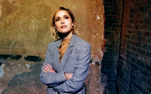 Stylish woman in a checkered blazer poses indoors with arms crossed, looking up against a textured wall with vintage tones.
