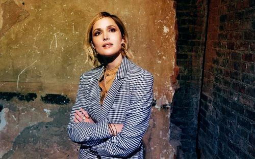 Stylish woman in a checkered blazer poses indoors with arms crossed, looking up against a textured wall with vintage tones.