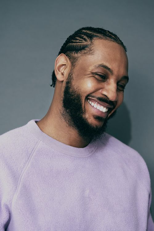 Portrait of a smiling adult male with braided hair and a beard wearing a lilac sweater against a gray background, captured in a close-up shot.