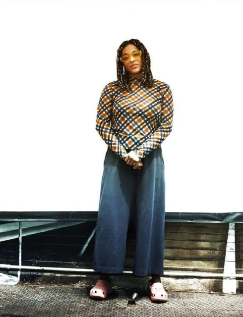 A woman in a plaid top and wide-leg pants stands against a wall outdoors, wearing translucent yellow glasses and pink clogs.