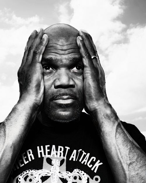 Monochrome portrait of a Black man with a bald head, his hands raised near his head, wearing a t-shirt, set against a cloudy sky background.