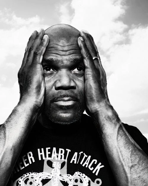 Monochrome portrait of a Black man with a bald head, his hands raised near his head, wearing a t-shirt, set against a cloudy sky background.