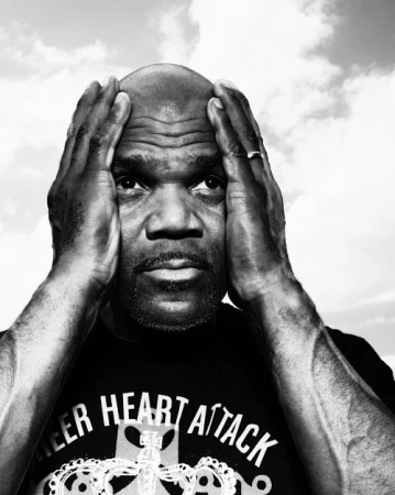 Monochrome portrait of a Black man with a bald head, his hands raised near his head, wearing a t-shirt, set against a cloudy sky background.
