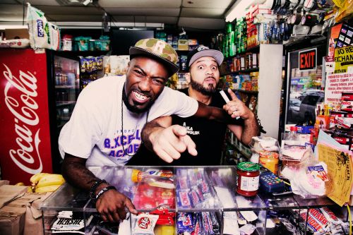 Two adult males pose behind a shop counter, with various items and a Coca-Cola fridge in the background, capturing an indoor setting.