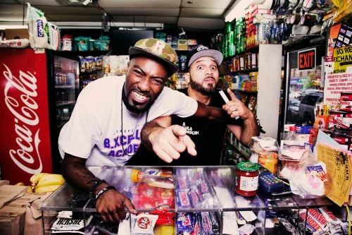 Two adult males pose behind a shop counter, with various items and a Coca-Cola fridge in the background, capturing an indoor setting.