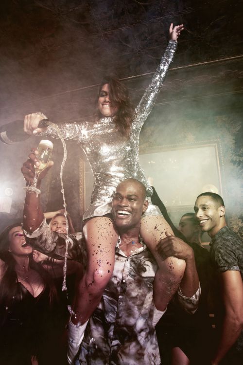 Woman in sequin dress sitting on shoulders of a smiling man while pouring champagne in a crowded celebratory party scene