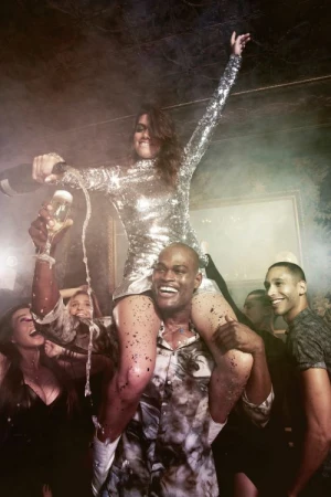 Woman in sequin dress sitting on shoulders of a smiling man while pouring champagne in a crowded celebratory party scene