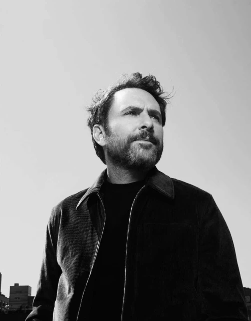 Black-and-white portrait of an adult male with beard and casual jacket against a clear sky and urban background. Lifestyle shot.
