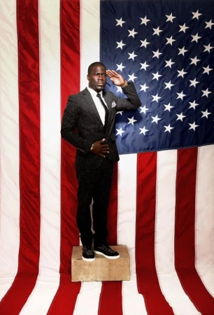 Portrait of adult male saluting in a grey suit and sneakers in front of a large US flag backdrop in an indoor photo studio.