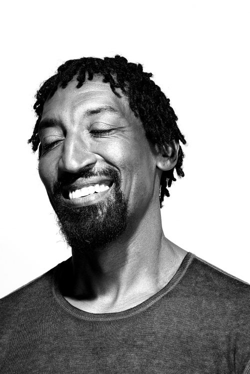 Close-up portrait of an African American man with dreadlocks, a beard, and a gray shirt, smiling with his eyes closed in a studio setting.