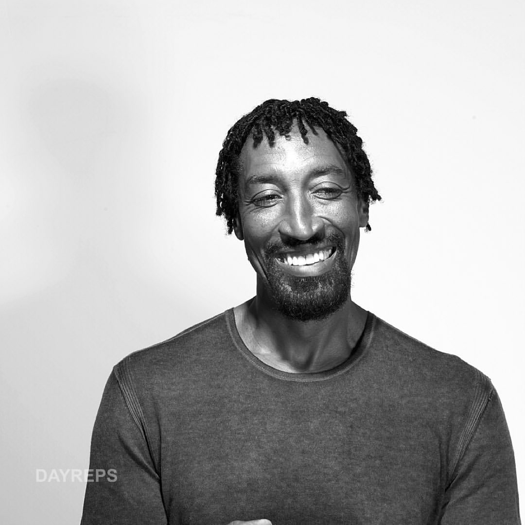 Black and white portrait of a smiling adult male with short dreadlocks wearing a crew neck shirt against a neutral background.