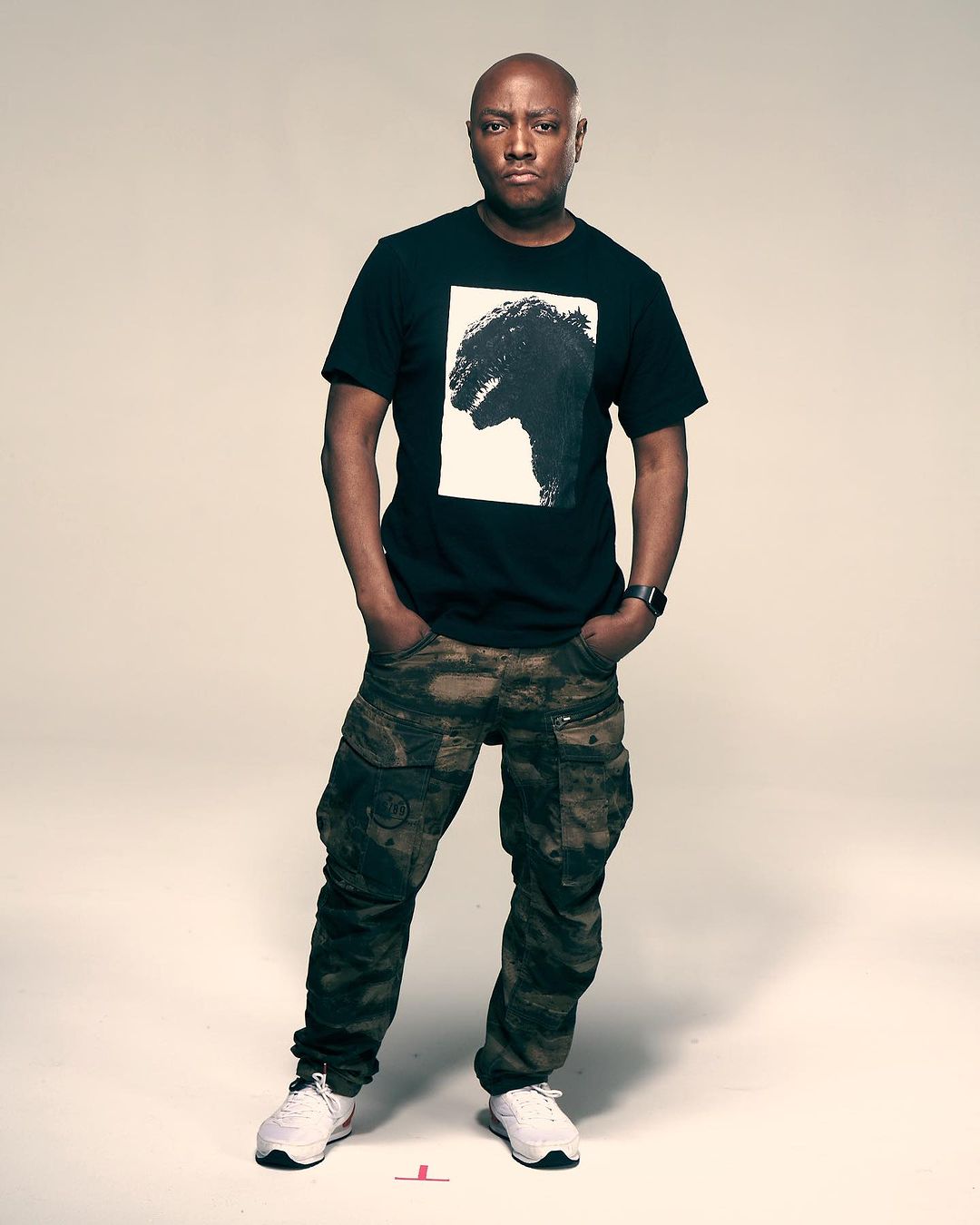 Full-length portrait of an adult male with a shaved head wearing a t-shirt with a graphic print, camouflage pants, and white sneakers.