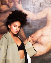 Portrait of a Black woman with voluminous afro hair wearing a muted green patterned coat and black lace bra standing against a painted classical art mural.