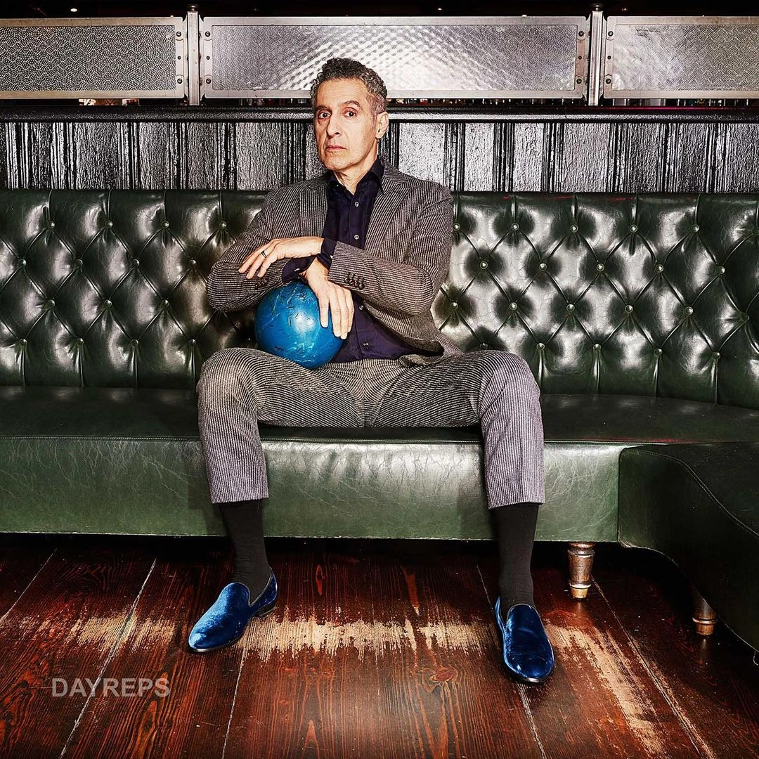 Man in a grey suit and blue velvet loafers sitting on a green tufted leather sofa holding a blue bowling ball, photographed in Los Angeles.