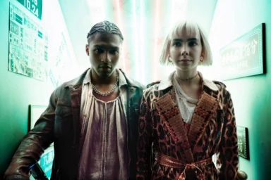 Two individuals posing in a narrow corridor with green walls, featuring a man in a leather jacket and a woman in a leopard print coat.