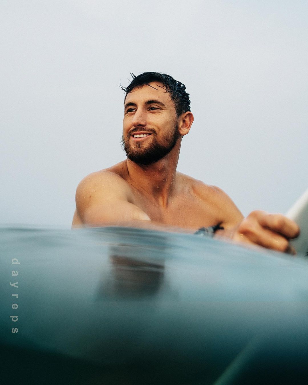 A man with short dark hair smiles in the water, holding a surfboard. The water is calm with a gray sky above, medium shot.