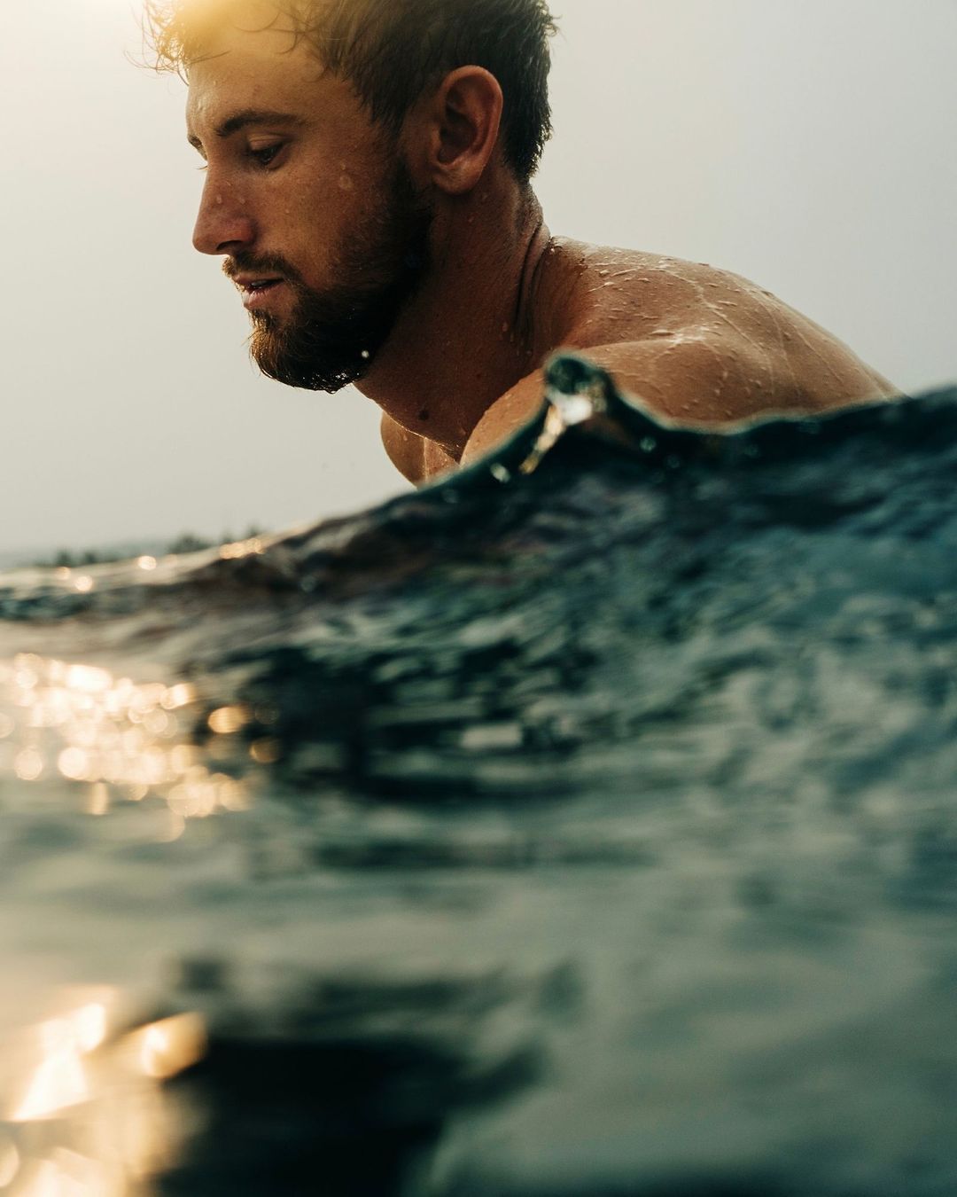 Adult male swims in ocean at golden hour, side profile with wet hair, looking downward with sunlight reflecting on water, low horizon.