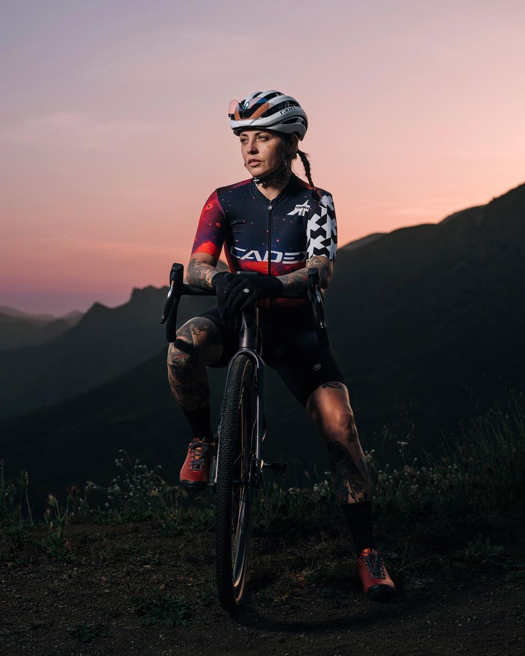 Female cyclist in galaxy-themed jersey pauses on a mountain trail with her bicycle, enjoying the dusk mountain views.
