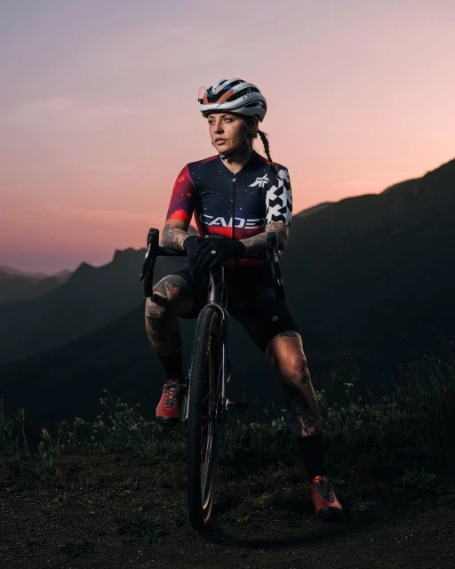 Female cyclist in galaxy-themed jersey pauses on a mountain trail with her bicycle, enjoying the dusk mountain views.