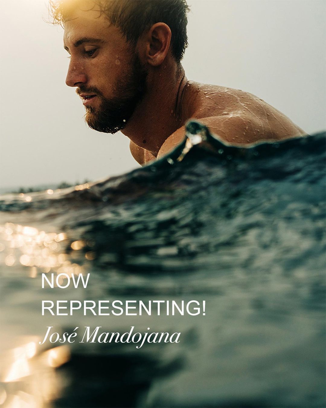 A man with a beard emerges from the sea, with visible waves. The photo captures the moment with water droplets on his skin.