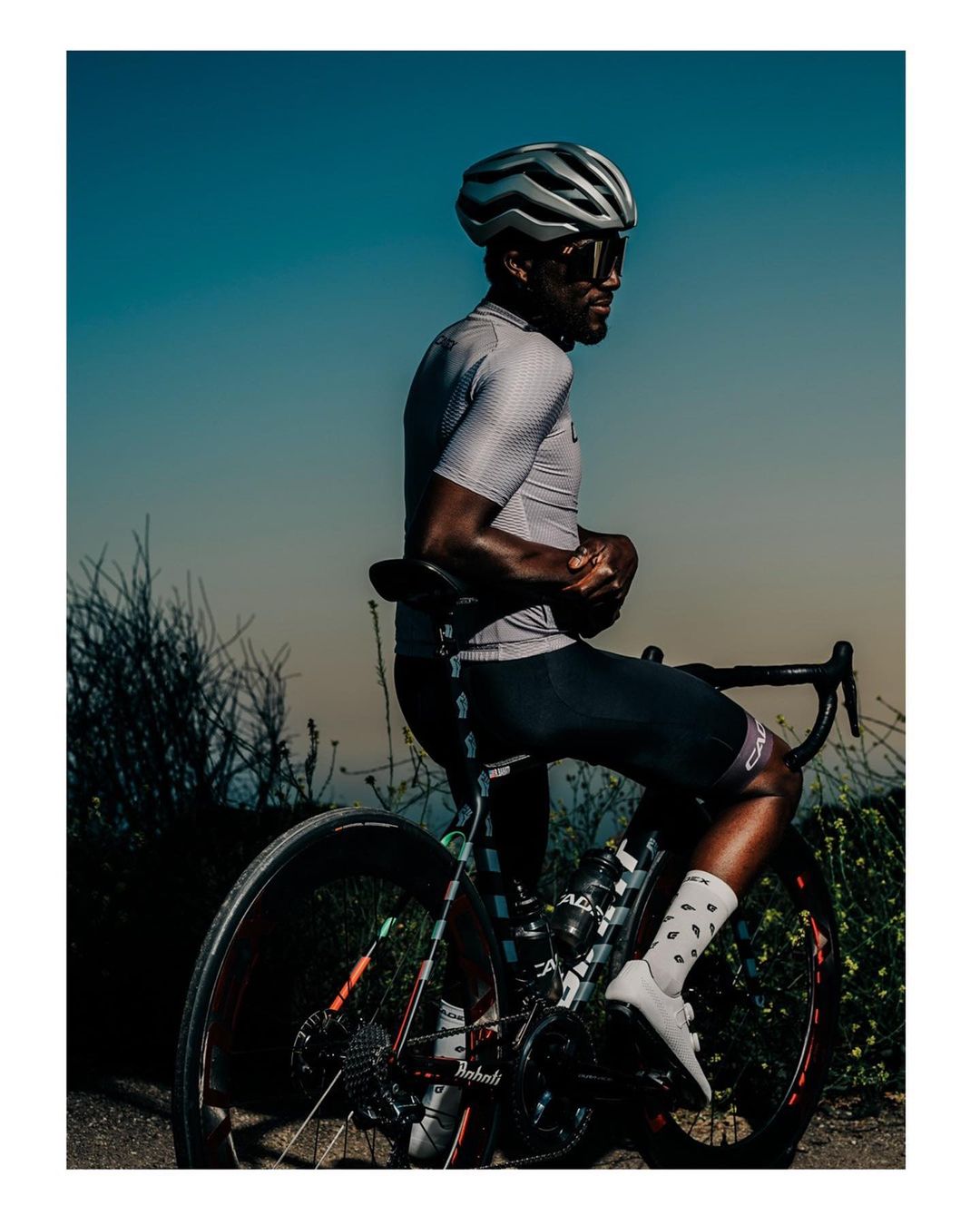 A cyclist wearing sunglasses and a helmet seated on a road bike outdoors, under a clear sky, with sparse vegetation in the background.
