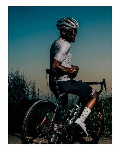 A cyclist wearing sunglasses and a helmet seated on a road bike outdoors, under a clear sky, with sparse vegetation in the background.