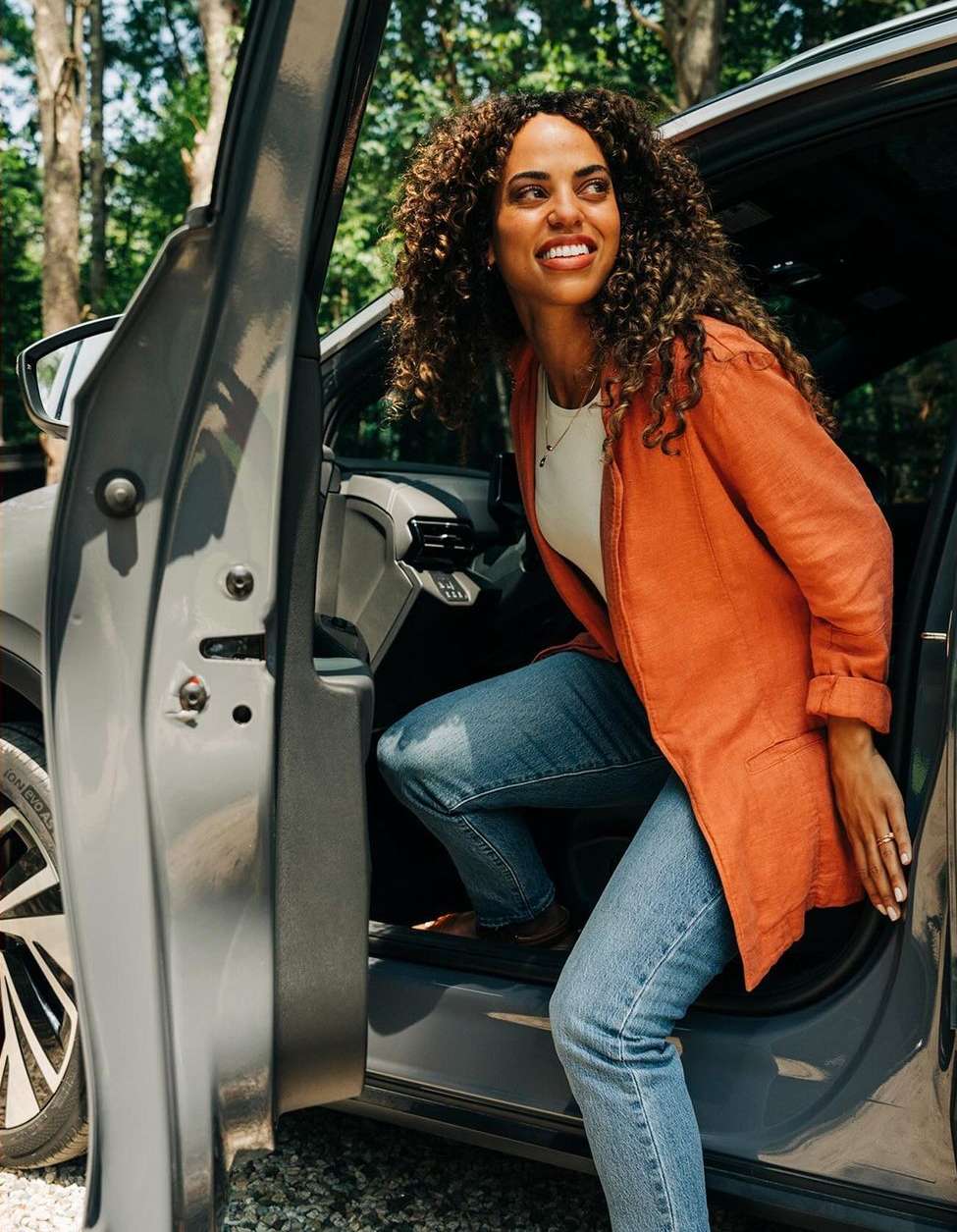 A brunette woman with curly hair in an orange jacket exits a gray car parked in a wooded area during the day.