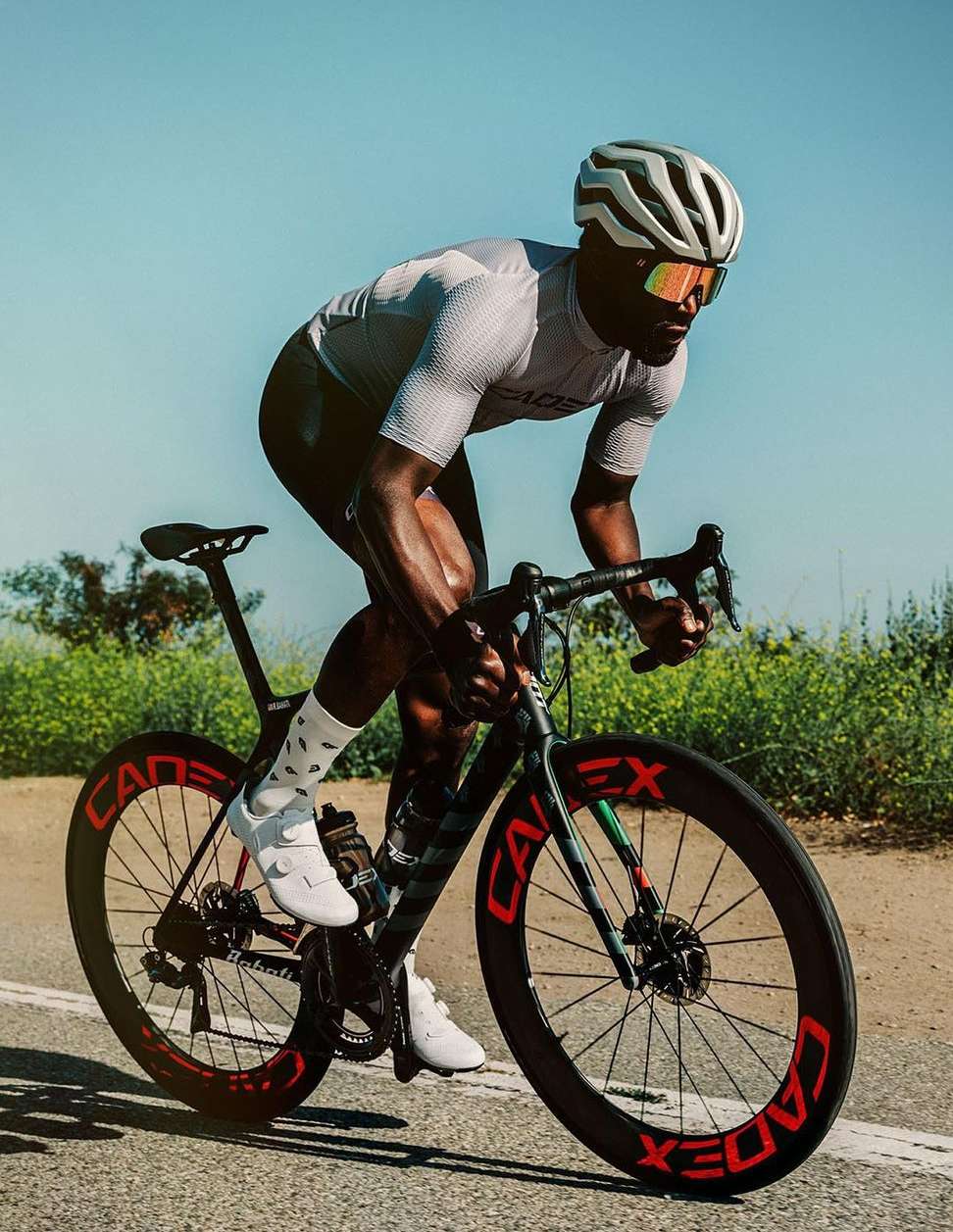 African American male cyclist in helmet and sunglasses riding black Cadex bike on asphalt road under bright sky, fitness and sport.