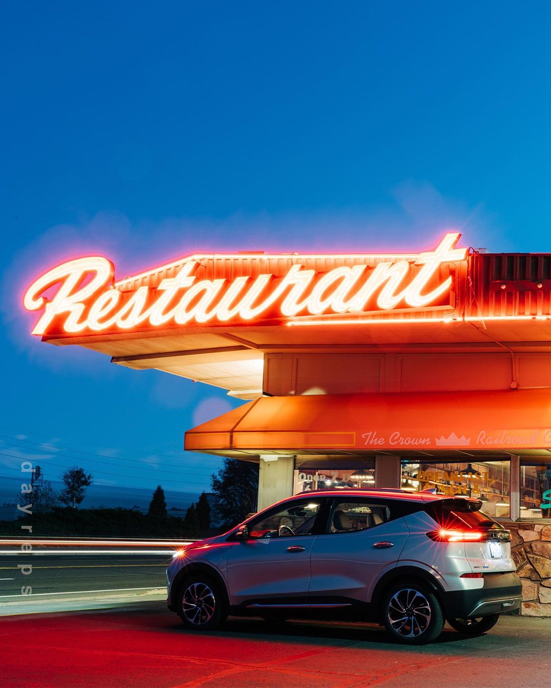 Silver electric vehicle parked in front of a vintage neon-lit restaurant at dusk, showcasing automotive design and retro roadside architecture.