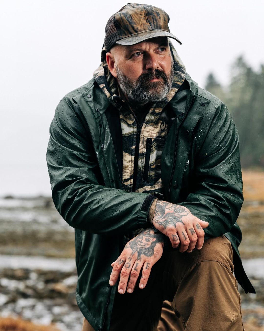 Man in a camouflage cap and green jacket kneeling in an outdoor setting. The background features a blurred landscape with trees.