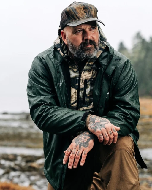 Man in a camouflage cap and green jacket kneeling in an outdoor setting. The background features a blurred landscape with trees.