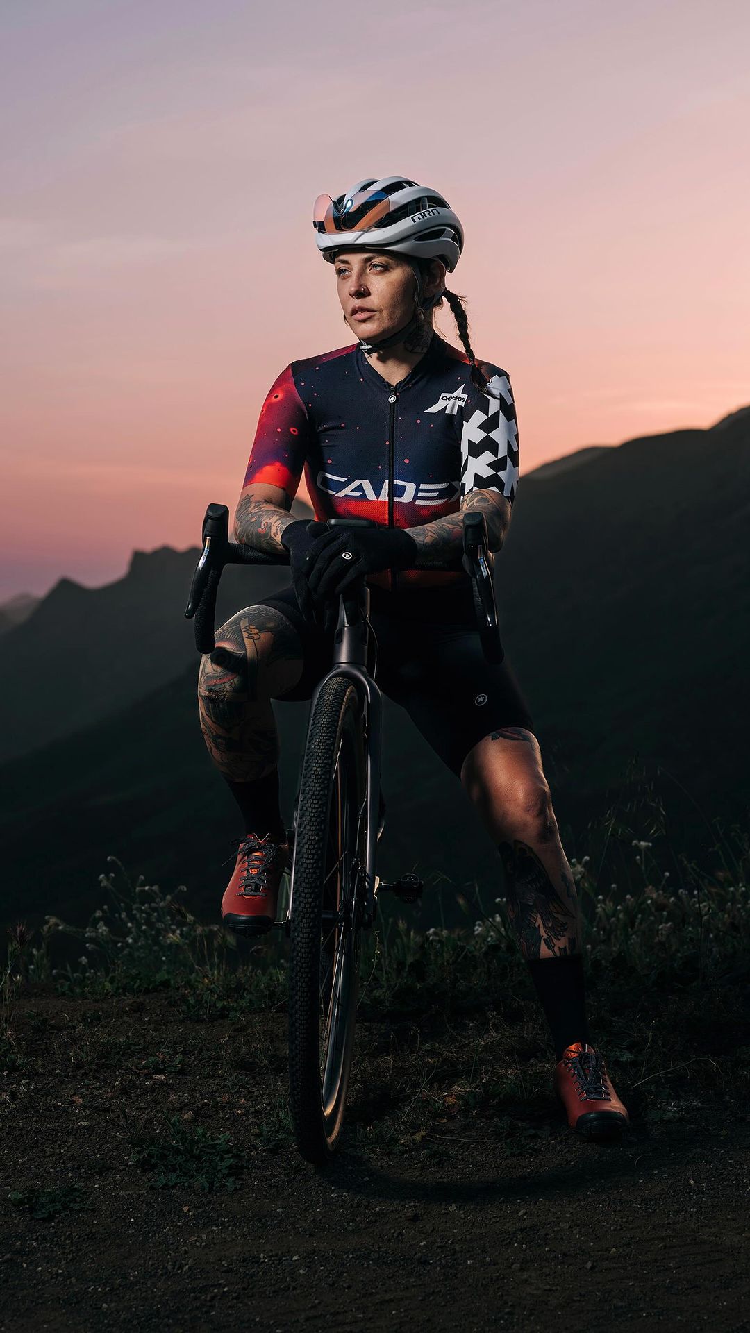 Woman cyclist in sportswear poses with her bicycle on a dirt path, set against mountains at dusk with orange and black sky.
