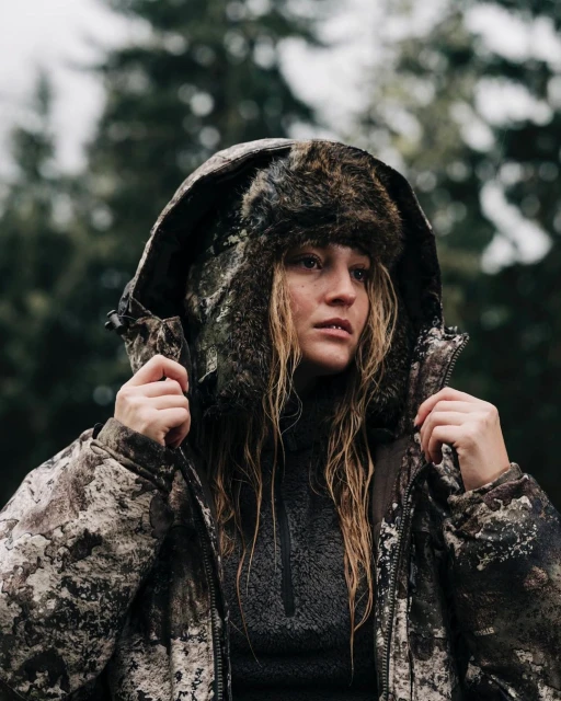 Adult woman in a hooded camouflage jacket and fur hat stands outdoors in a forest setting, glancing to the side with neutral expression.