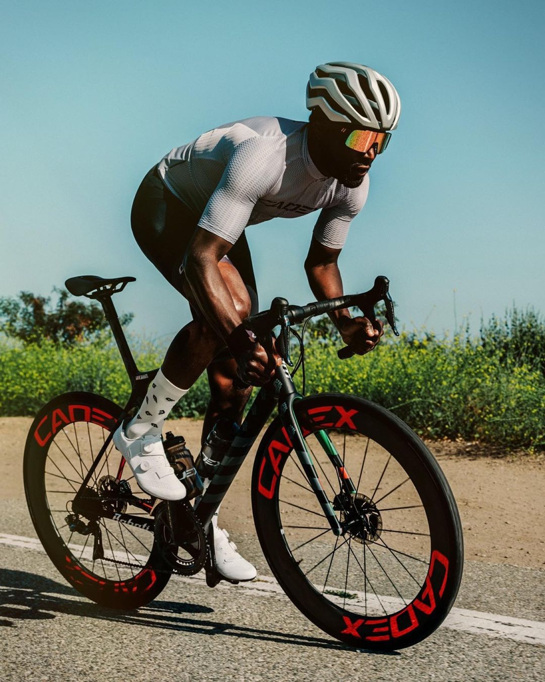 An adult male road cyclist wearing a helmet, sunglasses and jersey is riding a road bike on a paved road on a sunny day outdoors.