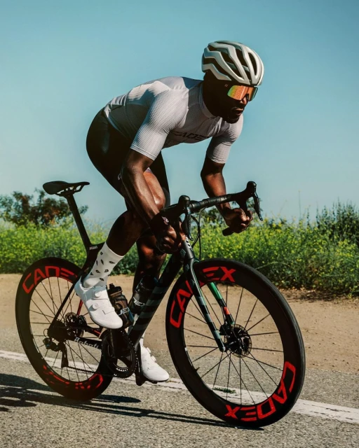 An adult male road cyclist wearing a helmet, sunglasses and jersey is riding a road bike on a paved road on a sunny day outdoors.