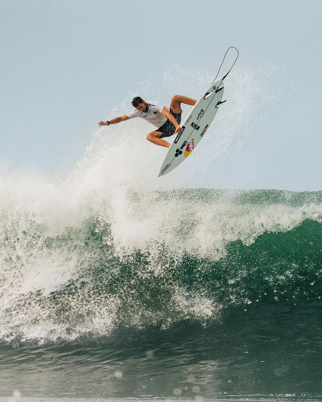 A surfer performs an aerial maneuver on a surfboard above a large wave, showcasing athleticism against a clear sky backdrop.