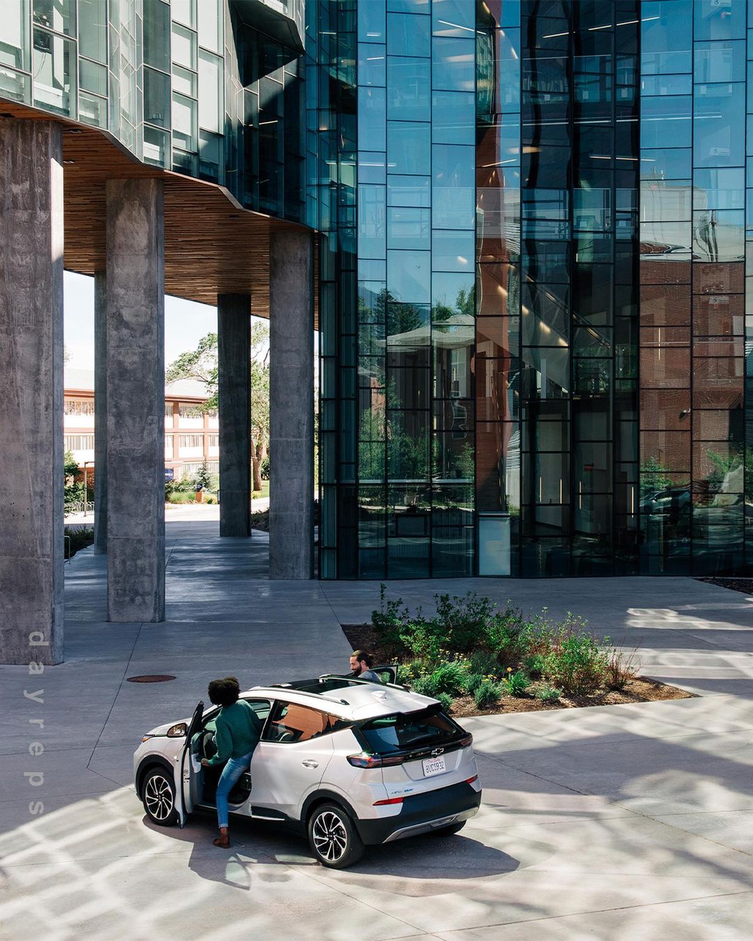 A white electric SUV is parked next to a modern glass building, with a woman standing near the open driver's side door on a sunny day.