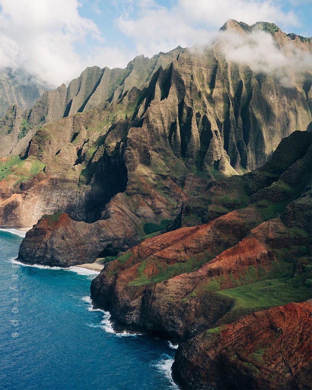 Aerial view of jagged, rocky mountains and cliffs along the coast of Kauai, Hawaii. Blue ocean water meets the cliffs below a cloudy sky.