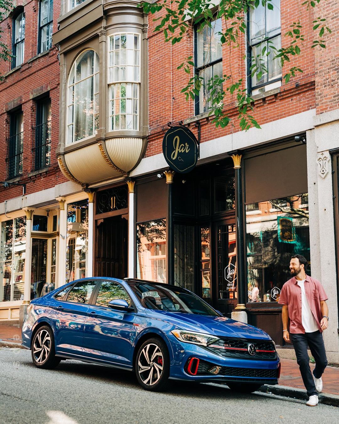 A blue Volkswagen Jetta car is parked on a city street, with a man walking next to it and a brick building in the background.