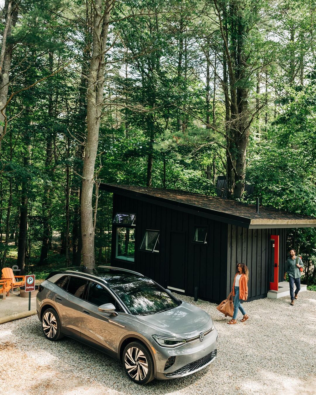 Silver Volkswagen ID.4 electric SUV parked near a modern black house in a wooded area with two adults walking toward the home.