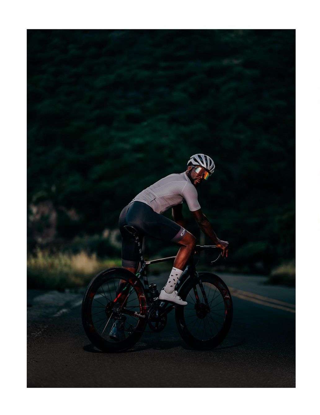 Man in cycling gear rides a road bike on a paved road at dusk, with dark green foliage in the background. He wears a helmet and sunglasses.