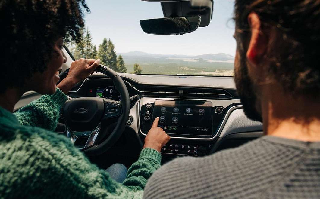 Two people inside a car with an infotainment system displaying various apps. Trees and mountains are visible through the windshield.