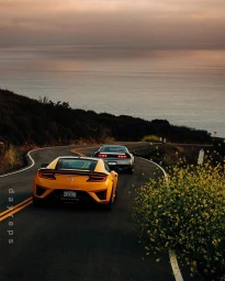 Rear view of a yellow sports car driving on a winding coastal road at sunset with the ocean in the background