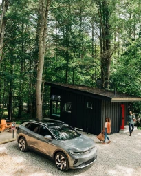 Silver electric SUV parked in a gravel clearing in front of a dark cabin surrounded by tall trees with a man and woman walking nearby.