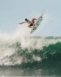 Surfer performing an aerial maneuver above a crashing green ocean wave under an overcast sky, captured by José Mandojana